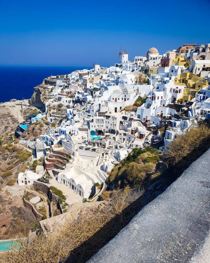 Santorini Greek Islands Greece A Complete Guide To The Santorini Public Transport System A hill in Santorini, Greece with heaps of white buildings going down the hill. There's a windmill in the distance and some blue water can be seen behind the hill "A Complete Guide To The Santorini Public Transport System"