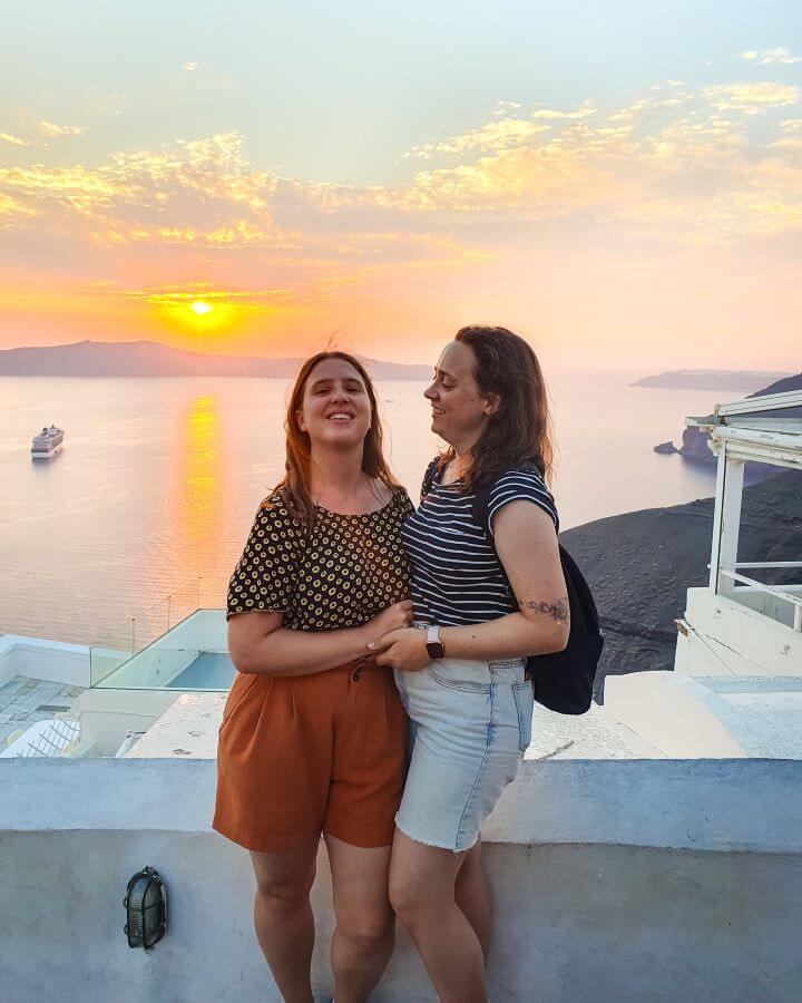 Fira Santorini Greek Islands Greece A Complete Guide To The Santorini Public Transport System Katherine and Krissie smiling and holding each standing in front of a barrier with the Santorini caldera in the Greek Islands behind them. In the water is a cruise ship and behind it all is a beautiful yellow and orange sunset "A Complete Guide To The Santorini Public Transport System"