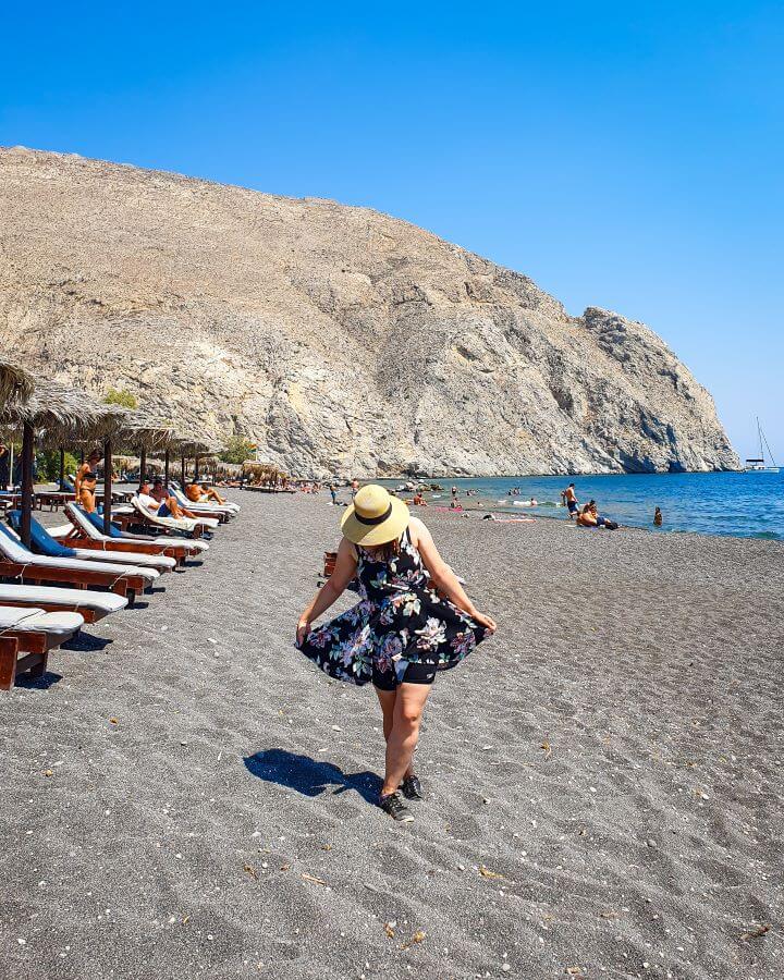 Black Sand Beach Santorini Greek Islands Greece A Complete Guide To The Santorini Public Transport System Katherine looking down and holding her dress on a black sand beach in Santorini, Greece. Behind her are sun chairs, the blue water and a huge brown hill behind it all "A Complete Guide To The Santorini Public Transport System"