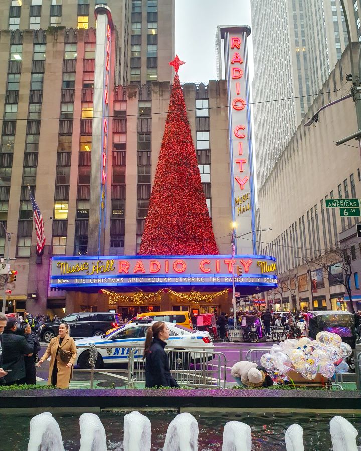 Radio City Music Hall New York City USA The Ultimate Guide for Musical Theatre Fans In New York Radio City Music Hall on 6th Avenue in New York City, USA. Radio City has a couple of big signs on the front in yellow and red lights and the big tree on top is red with a big red star on top. In front of it is a road with cars and lots of people around. In the very front of the photo is a fountain with some water spouts "The Ultimate Guide for Musical Theatre Fans In New York"