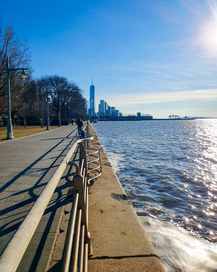 Hudson River New York City USA Non-Touristy Activities for Return Visitors to New York City Looking down along the Hudson River in New York City, USA. On the left is a path with a metal railing alongside it with some light poles and and trees along the side of the path. On the right is the blue water with the blue sky above it all and in the distance are some tall high rise buildings at the bottom of Manhattan "Non-Touristy Activities for Return Visitors to New York City"