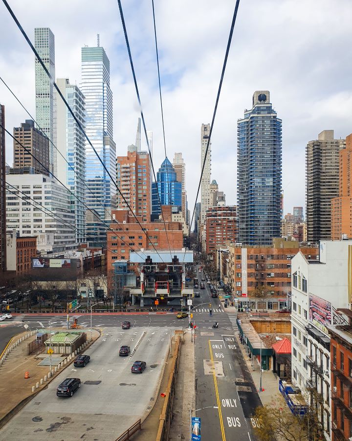 Roosevelt Island Tramway New York City USA Non-Touristy Activities for Return Visitors to New York City A view from the tramway that goes from Manhattan over to Roosevelt Island in New York City, USA. There's 8 wires coming from the top of the photo leading back to the building the tramway comes from. Around that building are the high rises of Manhattan, roads, cars and people walking around "Non-Touristy Activities for Return Visitors to New York City"