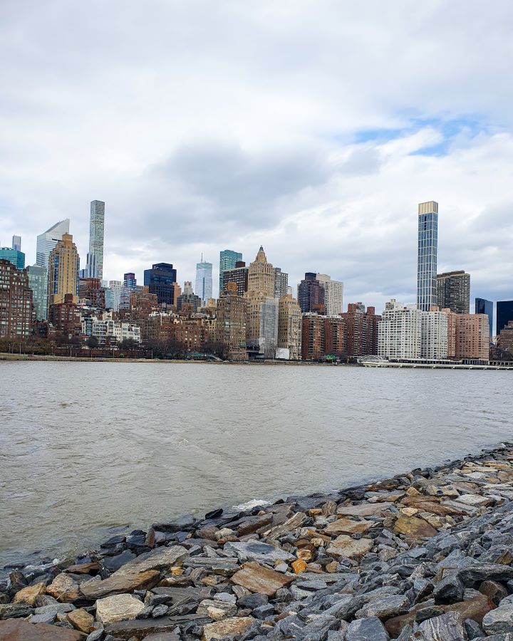Roosevelt Island New York City USA Non-Touristy Activities for Return Visitors to New York City A view back to Manhattan from Roosevelt Island New York City, USA. In the foreground are rocks on the bank of the island, with the brown river behind it. Beyond the river is Manhattan which has lots of high rises and the sky is really cloudy (it ended up snowing this day) "Non-Touristy Activities for Return Visitors to New York City"
