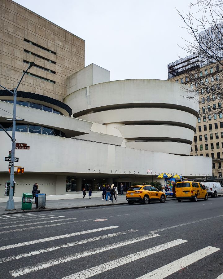 Guggenheim Museum New York City USA Non-Touristy Activities for Return Visitors to New York City The Guggenheim Museum in New York City, USA. In the foreground is the road with a couple of yellow taxis on it. Behind them is the museum which is a big cream coloured building that has a bowl shape on top with black stripes on it "Non-Touristy Activities for Return Visitors to New York City"