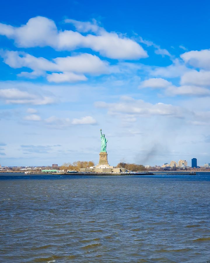 Statue of Liberty New York City USA Non-Touristy Activities for Return Visitors to New York City A view of the Statue of Liberty that's in the middle of the photo and is close enough to see that there's people on the island. In front of the statue is lots of water and behind you can see more plants and buildings on the land "Non-Touristy Activities for Return Visitors to New York City"