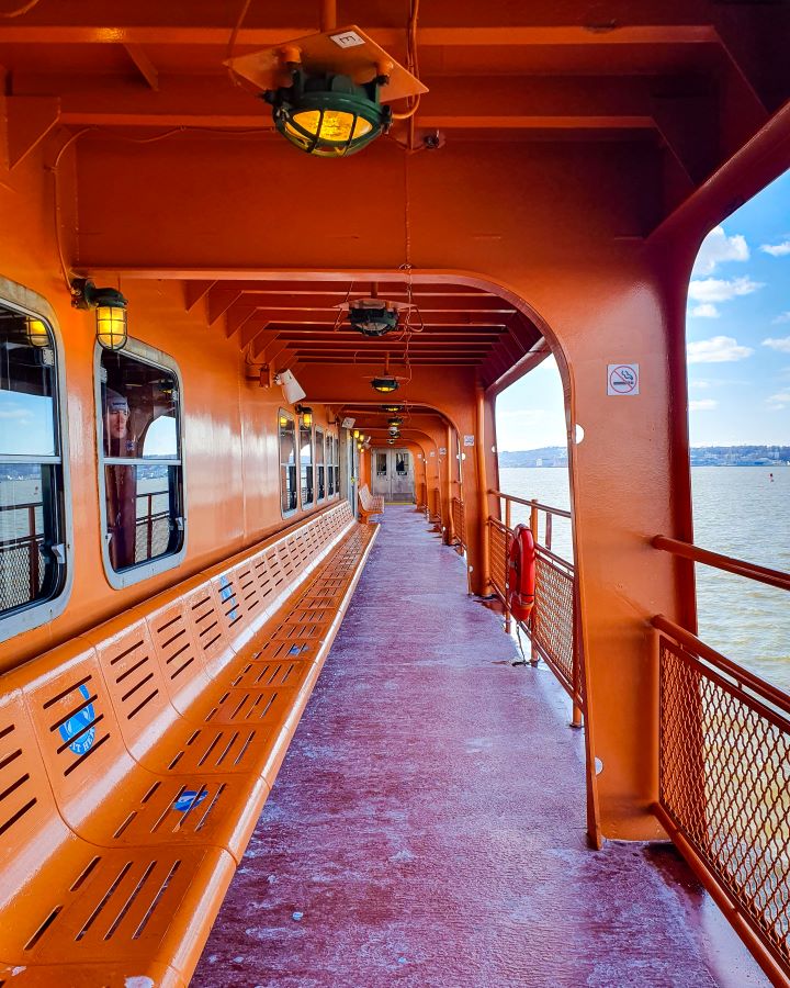 Staten Island Ferry New York City USA Non-Touristy Activities for Return Visitors to New York City The outdoor section of the Staten Island Ferry in New York City, USA. The whole thing is a bright orange colour and you can see the water on the right. On the left is outdoor seating and there's a walkway leading to a door inside the ferry. Over the walkway and seating is the orange roof with some lights on it too and there's no other people around "Non-Touristy Activities for Return Visitors to New York City"