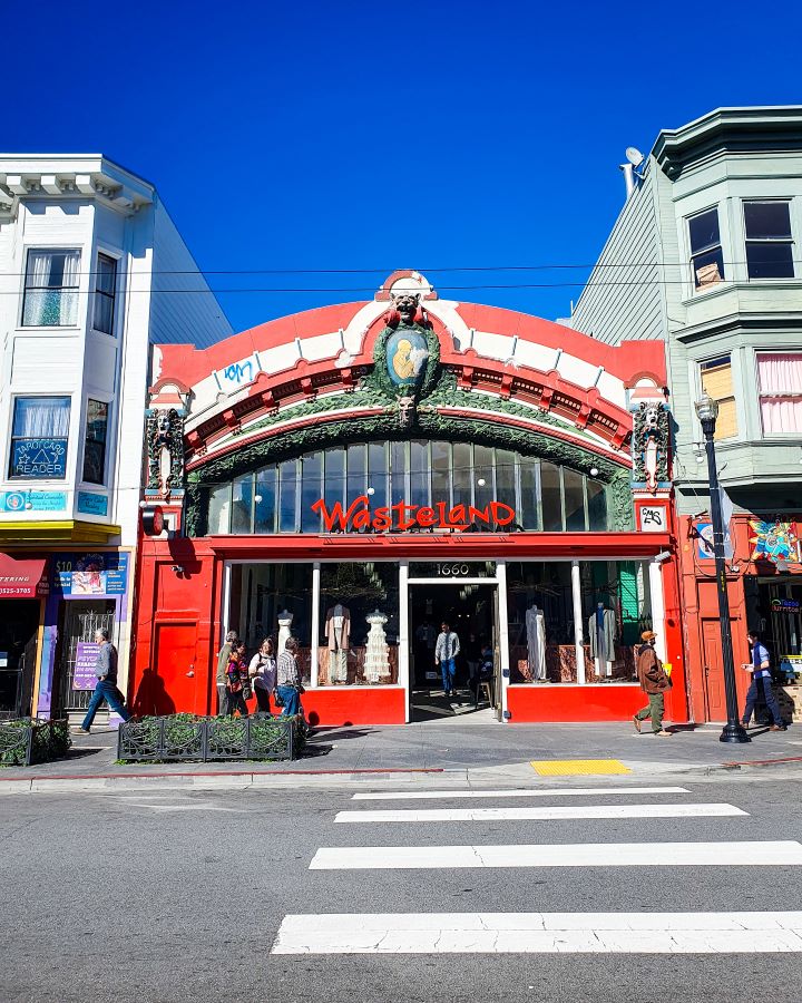 Haight Ashbury San Francisco USA How to See San Francisco on a Budget A red building in Haight Ashbury in San Francisco, USA. It says Wasteland in red writing on the front, and the building has an arched roof with long windows and clothes on mannequins out the front. There are people walking along the street and an empty road in the foreground. There's also buildings on either side of the Wasteland shop "How to See San Francisco on a Budget"