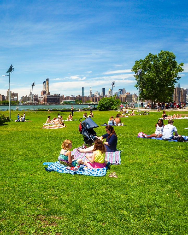Smorgasburg Williamsburg Brooklyn New York City USA Non-Touristy Activities for Return Visitors to New York City A large patch of green grass with people sitting on it and picnicking in the sun. Behind them in the river and across it is a view of Manhattan, with lots of high rise buildings. There's also trees and bushes on the grassy area and the sky is blue "Non-Touristy Activities for Return Visitors to New York City"