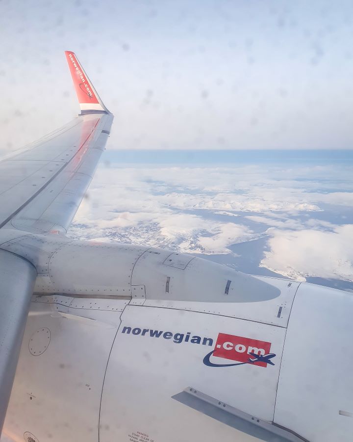 Northern Lights in Tromsø: Why It's the Best Place To Be A photo out the plane window flying over Norway looking at the wing with Norwegain.com on the engine. Below is a view of the snowy ground with water and clouds in between the snowy mountains. "Northern Lights in Tromsø: Why It's the Best Place To Be"
