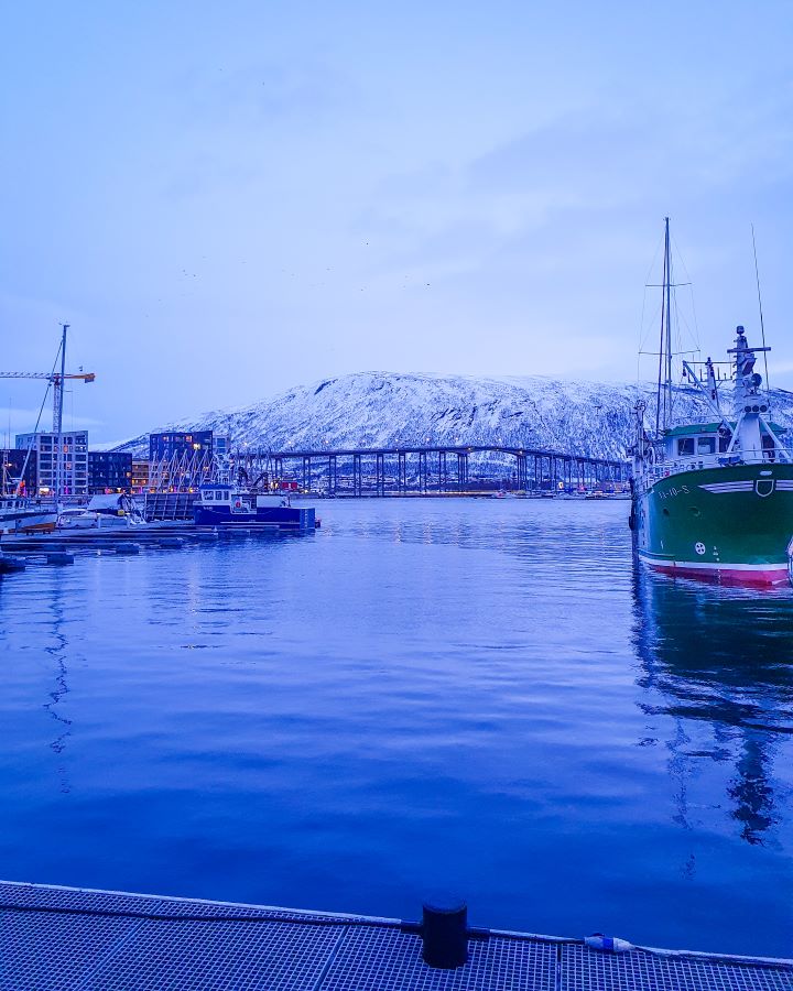 Best Things To Do in the City of Tromsø, Norway The Tromsø bridge in Tromsø, Norway. It's a big bridge with an arch in the middle. In front of it are boats sitting in the water, some buildings and behind it is a big snowy mountain. The photo is also very blue as the sun was going down. "Best Things To Do in the City of Tromsø, Norway"