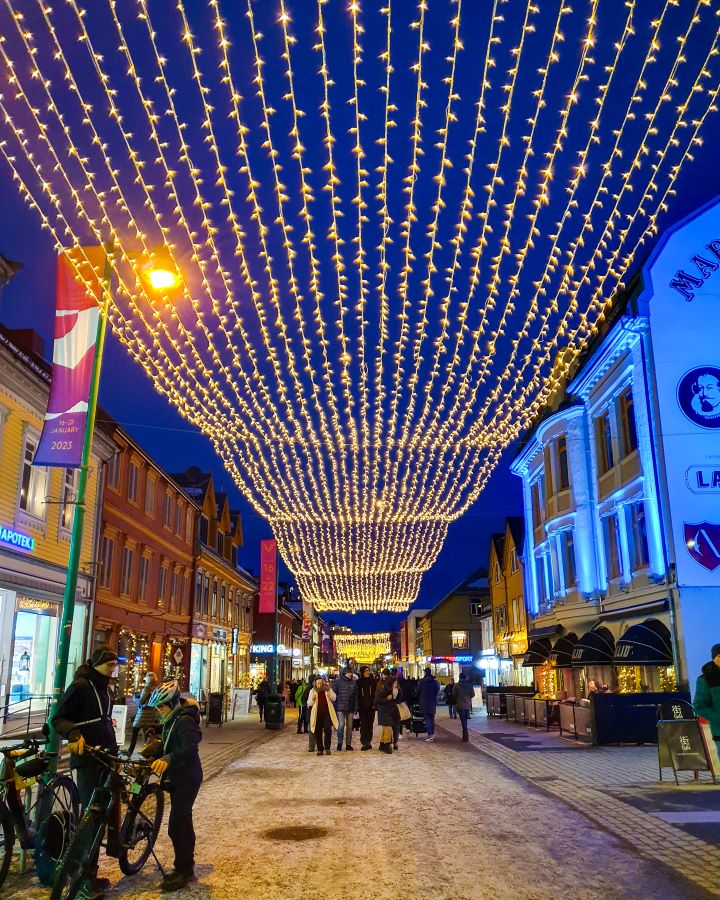 Northern Lights in Tromsø: Why It's the Best Place To Be Looking down the main street in Tromsø, Norway. There's shops on either side and fairy lights above the street running the whole way down. There's lots of people around and the sky is dark as the sun had gone down. "Northern Lights in Tromsø: Why It's the Best Place To Be"