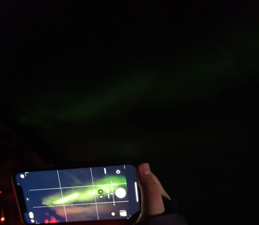 Northern Lights in Tromsø: Why It's the Best Place To Be A hand holding up a camera outside Tromsø, Norway, which has a very clear streak of green showing up on it. Behind the camera is a dark sky with a very very faint green streak. "Northern Lights in Tromsø: Why It's the Best Place To Be"