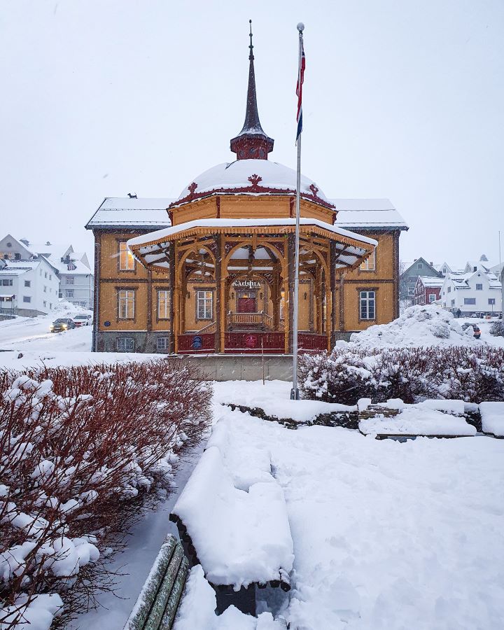 Northern Lights in Tromsø: Why It's the Best Place To Be Northern Lights in Tromsø: Why It's the Best Place To Be. A little yellow and red town hall in Tromsø, Norway with a bandstand out the front of the same colours with a big spire on top of it. In front of that is a flagpole and a little park with bushes and benches that's all covered in thick white snow and there's lots of houses behind it too.