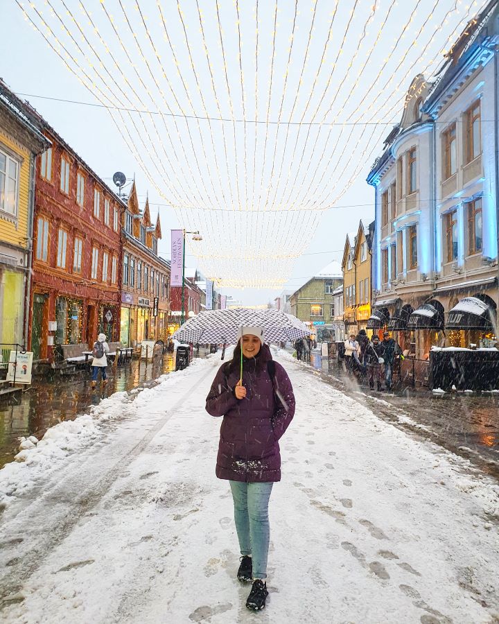 Best Things To Do in the City of Tromsø, Norway Krissie walking down the main street in Tromsø, Norway. She's holding an umbrella as it was snowing and there's snow on the ground. On either side of the street are coloured shops and there's fairy lights running all the way down the street above. "Best Things To Do in the City of Tromsø, Norway"