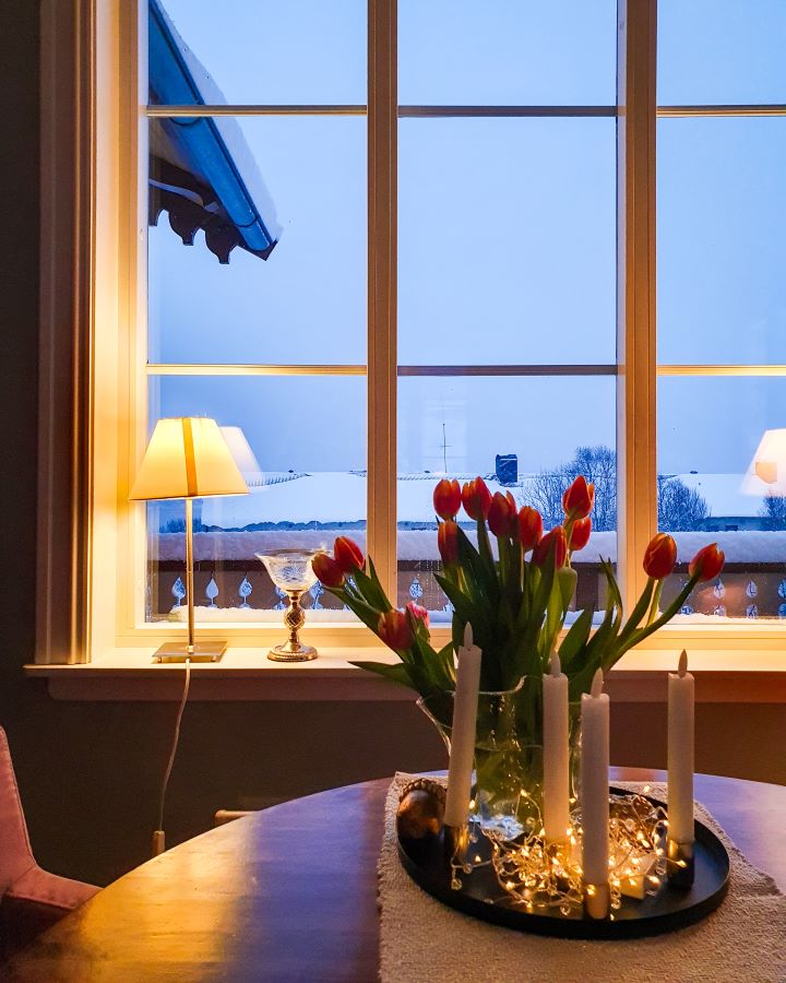 Northern Lights in Tromsø: Why It's the Best Place To Be Inside our Airbnb in Tromsø, Norway. There's a table with some fairy lights, fake candles and roses on it. Behind it is a lamp on the windowsill and outside the window is a snowy grey view. "Northern Lights in Tromsø: Why It's the Best Place To Be"