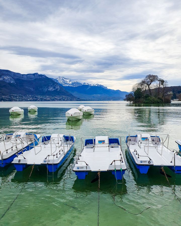 Four blue and white paddle boats sitting in the water in Lake Annecy, France. Behind the boats are some more white boats, a little island with trees, and huge hills behind it all with some snow on top. "How to Spend the Perfect Day in Annecy From Geneva"