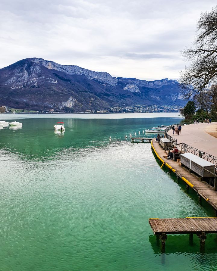 Lake Annecy in France. On the right side is a path with some little wooden jetties jutting out. There's also people walking along, trees and big hills in the background. "How to Spend the Perfect Day in Annecy From Geneva"