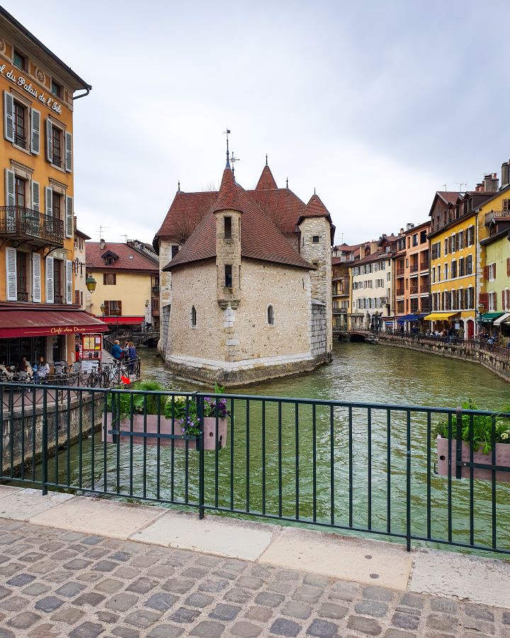 A view of the castle on Palais de I'Île in Annecy, France. In the foreground is the cobblestone of the bridge with a metal railing. Beyond that is the canal with Palais de I'Île in the middle which is a small castle looking building with a red pointed roof. The canal splits around the castle and on either side are coloured buildings with restaurants with outdoor seating. There's also flower boxes hanging off the bridge. "How to Spend the Perfect Day in Annecy From Geneva"
