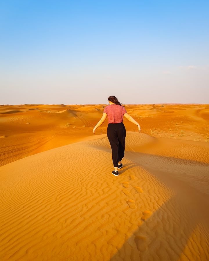Krissie standing on a hill of sand in the Arabian desert, UAE. She's walking away from the cameras and behind her are more hills of sand with a clear blue sky above it all. "Dubai Desert Safari Experience: Detailed Get Your Guide Review"