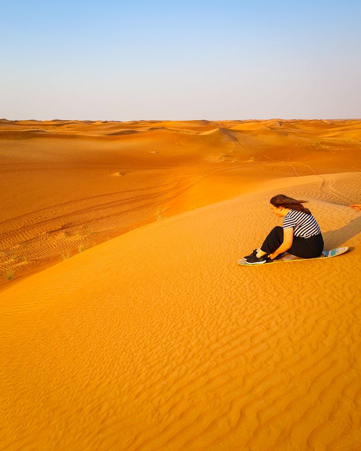 Katherine sitting on a sandboard which looks like a skateboard without wheels on the right side of the photo about to go down a sand hill. All around her is mountains of sand and a clear blue sky above it all. "Dubai Desert Safari Experience: Detailed Get Your Guide Review"
