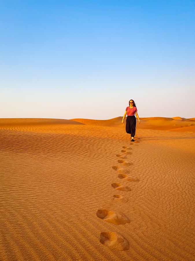 Krissie walking towards the camera with some footprints walking away from the camera in the Arabian Desert, UAE. All around her are hills of sand and the sky above is clear and blue. "Dubai Desert Safari Experience: Detailed Get Your Guide Review"