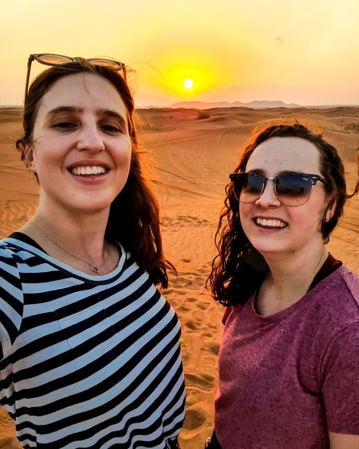 Dubai Desert Safari Experience: Detailed Get Your Guide Review. Katherine and Krissie smiling and looking at the camera with the Arabian Desert behind them and a bright yellow setting sun in the distance.