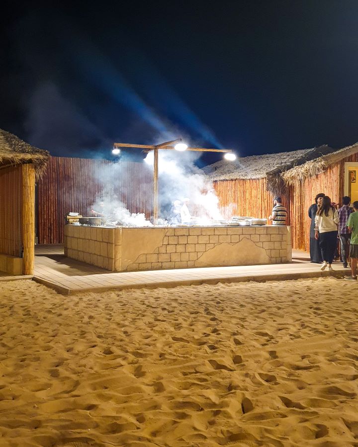 A brick barbeque with smoke coming off it at the dinner camp in the Arabian desert, UAE. Around it are little huts and people walking around with sand on the ground. "Dubai Desert Safari Experience: Detailed Get Your Guide Review"