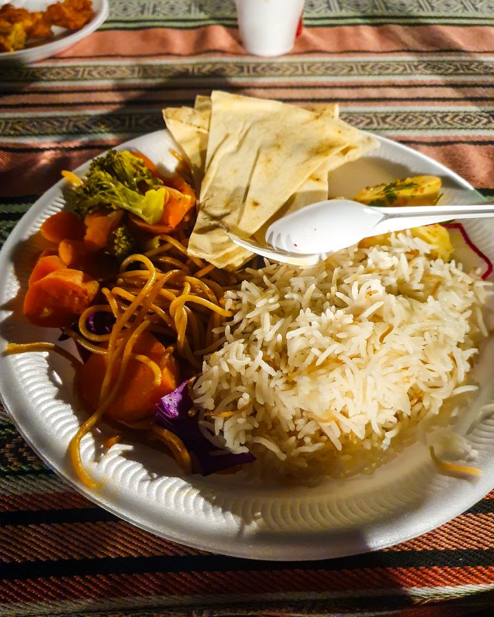 A white plate on a table with a stripy table cloth under it. On the plate are some rice, noodles, vegetables and bits of pita bread. "Dubai Desert Safari Experience: Detailed Get Your Guide Review"