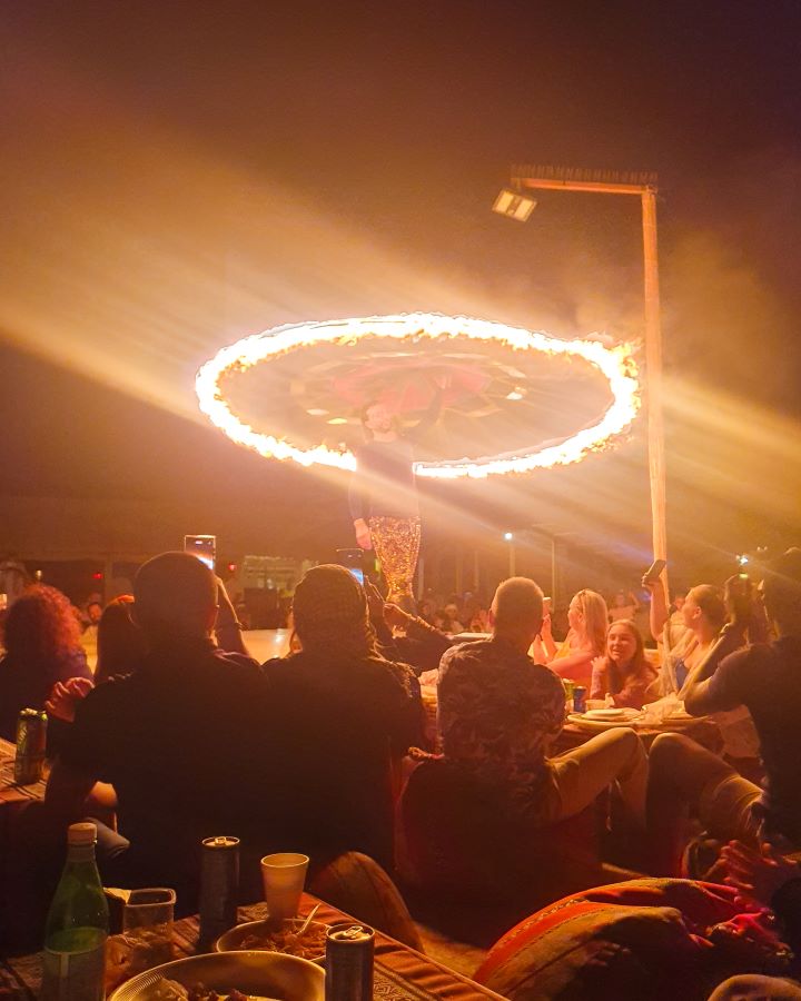 A man on a stage in the Arabian desert, UAE, with a flaming rope above him that's in a circle above his head. There's people sitting on the ground around him looking up at him. "Dubai Desert Safari Experience: Detailed Get Your Guide Review"