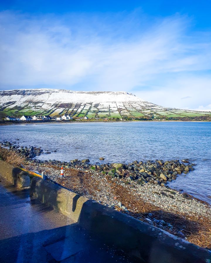 A view out the window of a bus in Northern Ireland. In the foreground is a rocky little beach with some water and a big green hill behind it with snow on it. "Giant's Causeway from Belfast: Get Your Guide Review"