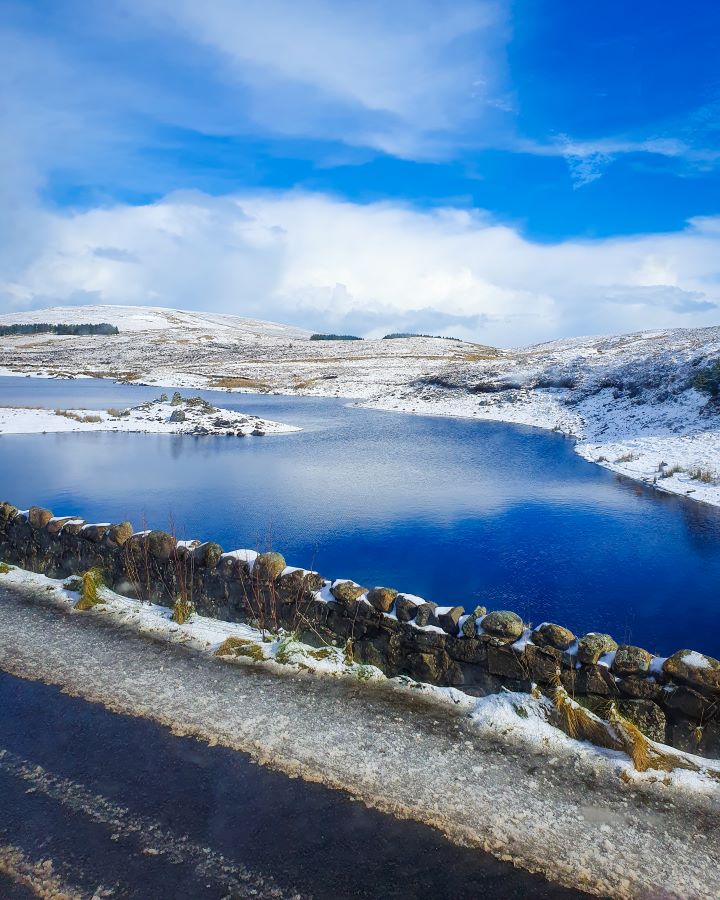 A view out the window in Northern Ireland. In the foreground is the road with a little rock barrier with a blue lake behind it. Around the lake are snowy hills with bushes on them. "Giant's Causeway from Belfast: Get Your Guide Review"