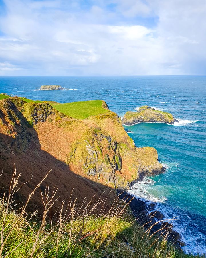 A view over an orange cliff with green grass on it in Northern Ireland. There's a couple of little islands off the mainland too and there's some green grass close to the camera. The water is very blue and the sky above is cloudy. "Giant's Causeway from Belfast: Get Your Guide Review"