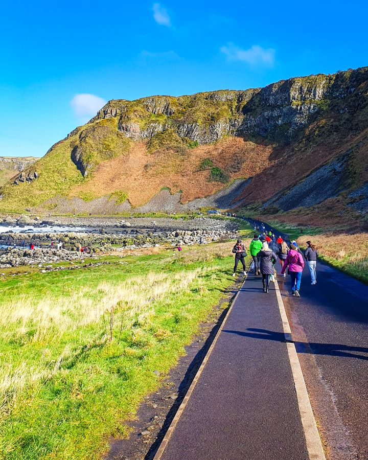 A road down to the Giant's Causeway in Northern Ireland. There's people in winter clothes walking down the road and behind it is a big green and orange hill and on the left are some rocks and water. "Giant's Causeway from Belfast: Get Your Guide Review"