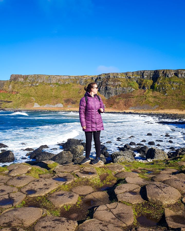 Krissie in a purple coat standing on some hexagonal rocks at the Giant's Causeway in Northern Ireland. Behind her is some blue water with black rocks and behind that is a huge orange and green cliff with blue sky above it. "Giant's Causeway from Belfast: Get Your Guide Review"