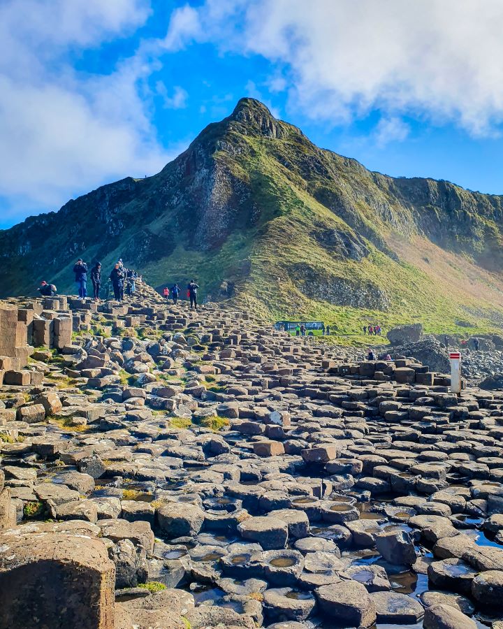 A bunch of hexagonal rocks with little pools of water in the middle of them with a big pointy green hill behind them. There's also people walking on the rocks and a cloudy sky above at the Giant's Causeway, Northern Ireland. "Giant's Causeway from Belfast: Get Your Guide Review"