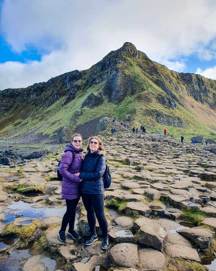 Giant's Causeway from Belfast: Get Your Guide Review. Krissie and Katherine wearing puffer jackets and hugging while standing in front of a big pointed green hill at the Giant's Causeway in Northern Ireland. Under them are lots of hexagonal rocks and there's people behind them on the rocks too.