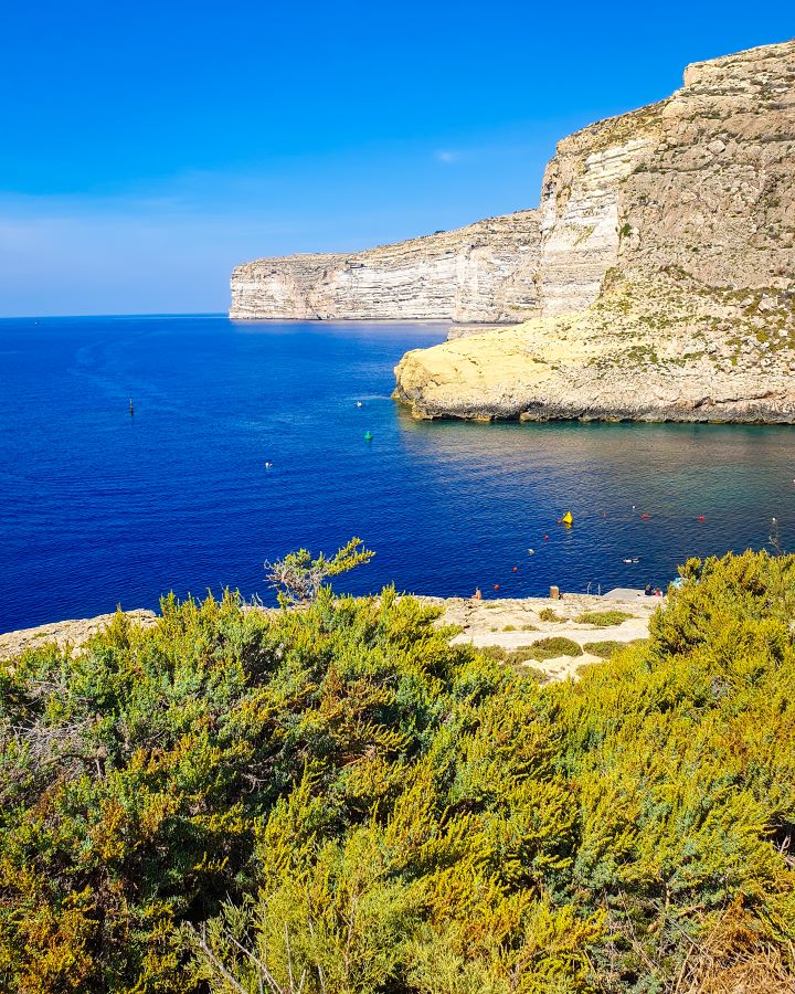 Xlendi Gozo Malta Explore Gozo and the Blue Lagoon in Malta: Detailed Get Your Guide Review A big white cliff on the right with blue water on the left in Xlendi, Gozo, Malta. In the foreground are lots of green bushes and there's a blue sky overhead. "Explore Gozo and the Blue Lagoon in Malta: Detailed Get Your Guide Review"