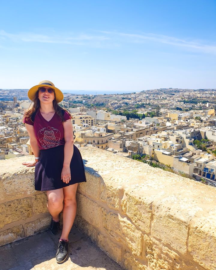 Victoria Citadel Malta Explore Gozo and the Blue Lagoon in Malta: Detailed Get Your Guide Review Explore Gozo and the Blue Lagoon in Malta: Detailed Get Your Guide Review. Katherine looking at the camera and leaning on a sandstone barrier in the Citadel in Victoria, Gozo, Malta. Behind her is a view over Gozo with lots of sandstone buildings, trees and a view of the water in the background.