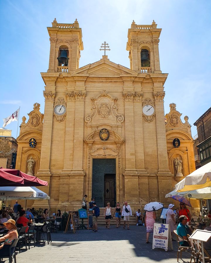 Victoria Malta Explore Gozo and the Blue Lagoon in Malta: Detailed Get Your Guide Review The big sandstone front of St. George’s Basilica in Victoria, Gozo, Malta. There's two bell towers on either side with little white clocks underneath. In the middle there's a big black doorway and stairs leading down to the street. There's heaps of people around with umbrellas on the street with people sitting down and eating under them. "Explore Gozo and the Blue Lagoon in Malta: Detailed Get Your Guide Review"