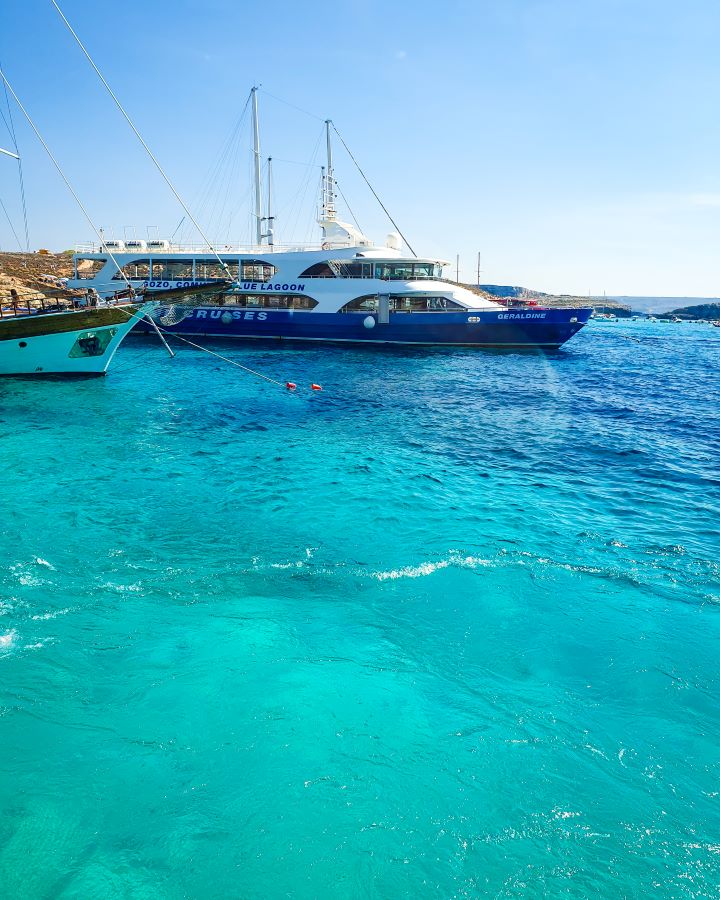 Blue Lagoon Comino Malta Explore Gozo and the Blue Lagoon in Malta: Detailed Get Your Guide Review Clear light blue water in the Blue Lagoon, Comino, Malta. In the background are a couple of boats with tall masts and on the left you can see a bit of brown land. "Explore Gozo and the Blue Lagoon in Malta: Detailed Get Your Guide Review"