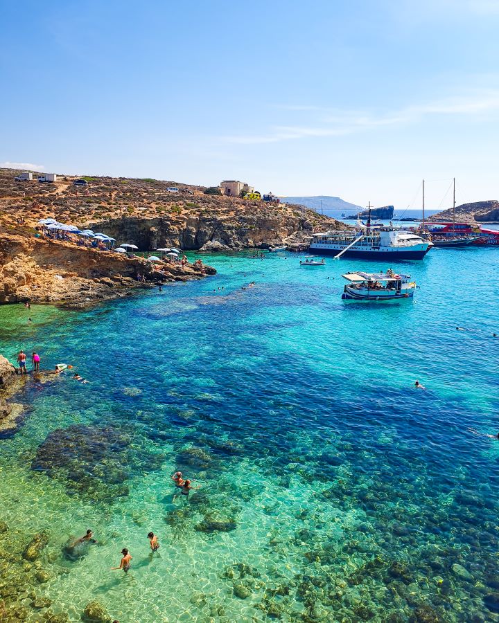 Blue Lagoon Comino Malta Explore Gozo and the Blue Lagoon in Malta: Detailed Get Your Guide Review In the foreground is light and dark blue clear water with people swimming in it. On the left is orange land with some blue umbrellas on it and on the right are some boats in the Blue Lagoon, Comino, Malta. "Explore Gozo and the Blue Lagoon in Malta: Detailed Get Your Guide Review"