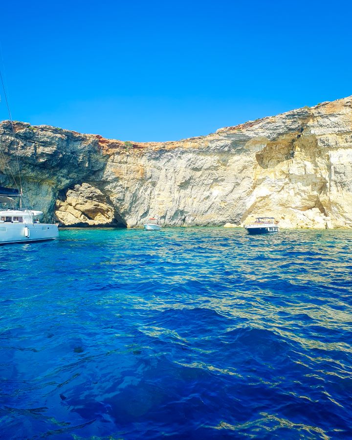 Blue Lagoon Comino Malta Explore Gozo and the Blue Lagoon in Malta: Detailed Get Your Guide Review In the foreground is clear blue water with some brown dots in the bottom left corner which are jellyfish in Malta. There's some boats on the water and behind them are some yellow cliffs with a little archway in the rock. "Explore Gozo and the Blue Lagoon in Malta: Detailed Get Your Guide Review"