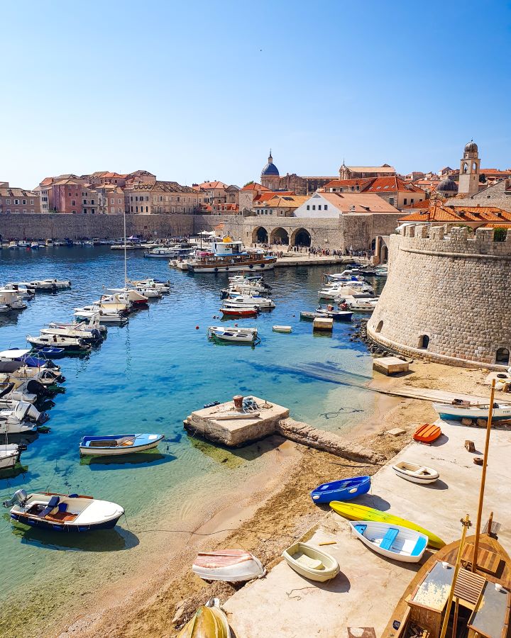 How to Spend Three Days in Dubrovnik: Best Itinerary For a First Visit. A view of Dubrovnik from outside the walls. In the foreground are little white boats in the clear blue water. Behind them are the sand coloured walls around the old town. Behind the walls are the city buildings with orange rooves and a clear blue sky above it all.