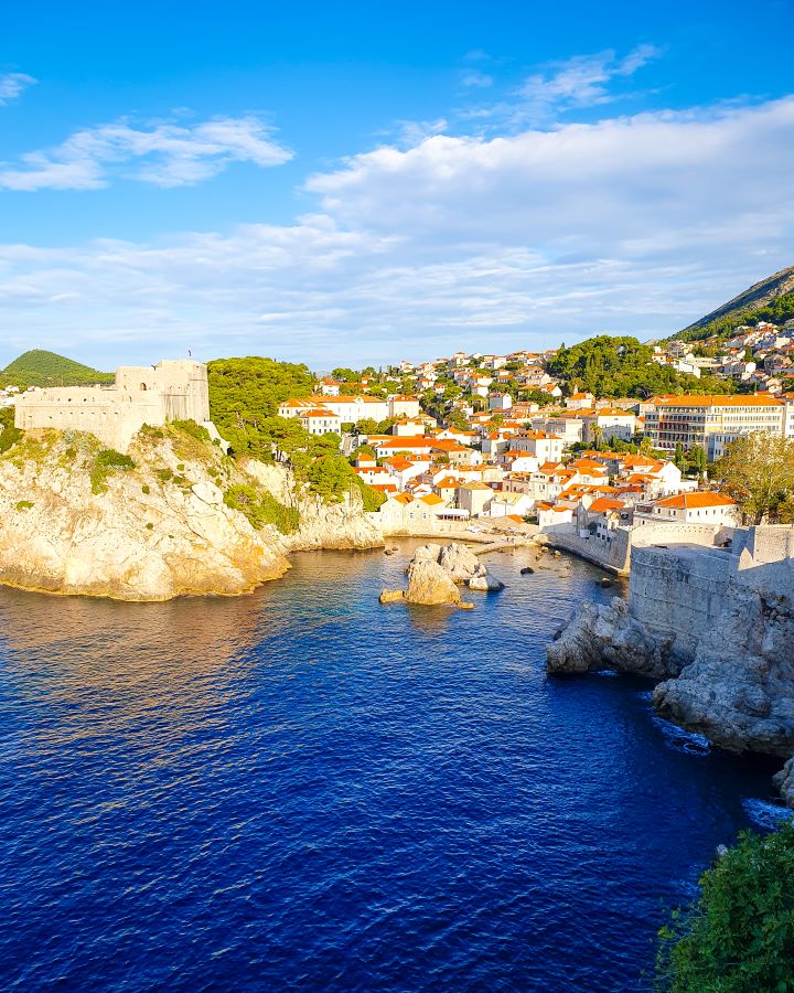 In the foreground is blue water with Dubrovnik old town on the right. On the left is the sand coloured Fort Lovrijenac on a hill and in between are more white buildings with orange rooves in Croatia. "How to Spend Three Days in Dubrovnik: Best Itinerary For a First Visit"