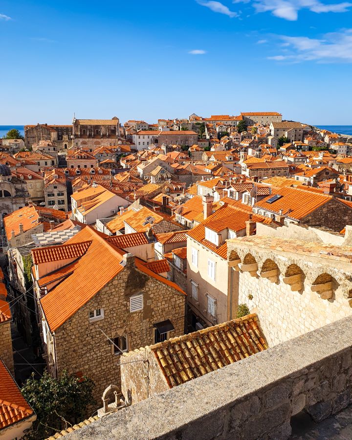 A view down on Dubrovnik old town in Croatia. The buildings have orange rooves and behind you can see a bit of the ocean. Above is also a blue sky. "How to Spend Three Days in Dubrovnik: Best Itinerary For a First Visit"