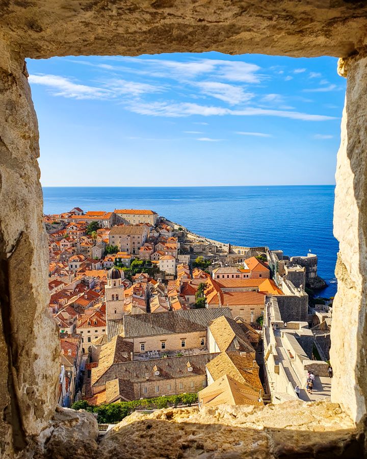 An incredible view out a stone window which frames the photo. Outside is a view down to Dubrovnik, Croatia with its orange rooves and clear blue water outside the walls. "How to Spend Three Days in Dubrovnik: Best Itinerary For a First Visit"