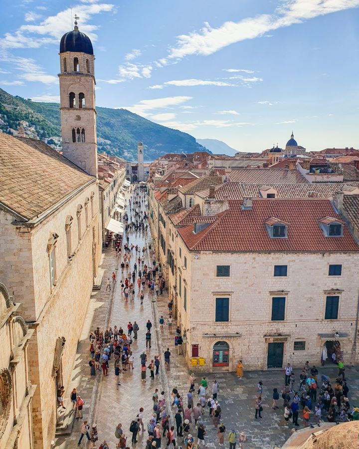 A view down on Stradun which is a long street in Dubrovnik, Croatia. There's lots of people walking around and on either side of the street are more sand coloured buildings with orange rooves and on the left is a church tower with a black dome on top. Behind the town are some green hills. "How to Spend Three Days in Dubrovnik: Best Itinerary For a First Visit"