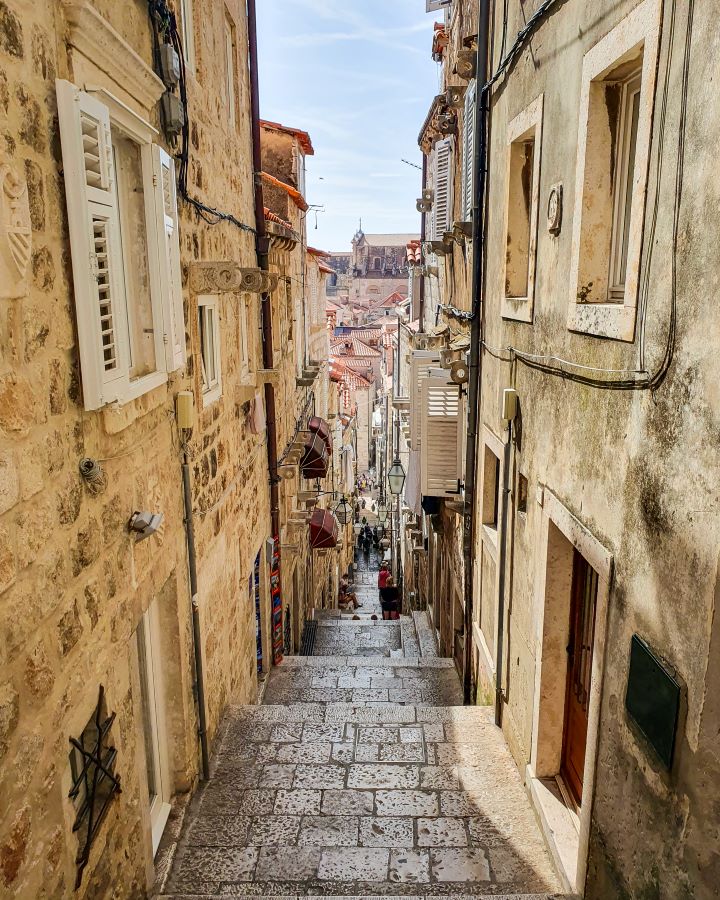 A view down a narrow street in Dubrovnik, Croatia. The buildings on either side are sand coloured and have white windows, and the path is made of white bricks. In the distance you can see more buildings with orange rooves in Dubrovnik, Croatia. "How to Spend Three Days in Dubrovnik: Best Itinerary For a First Visit"