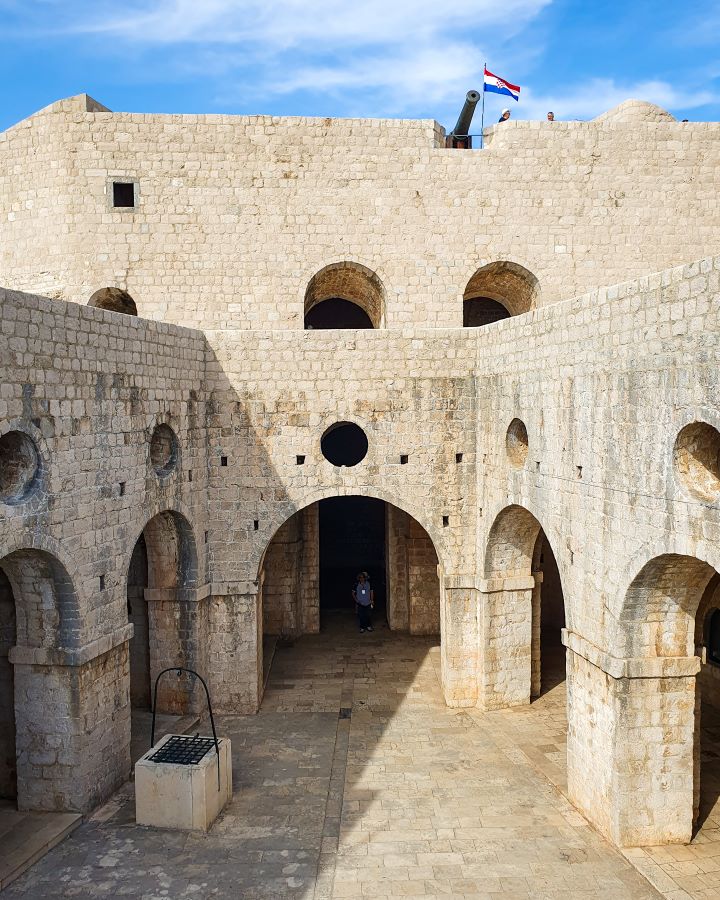 Inside Fort Lovrijenac in Croatia. There's a forecourt with a little well and above that are archways with circles in the walls above them and a canon up the top with the Croatian flag. The fort is made of sand coloured stone and above it is a blue sky with clouds. "How to Spend Three Days in Dubrovnik: Best Itinerary For a First Visit"