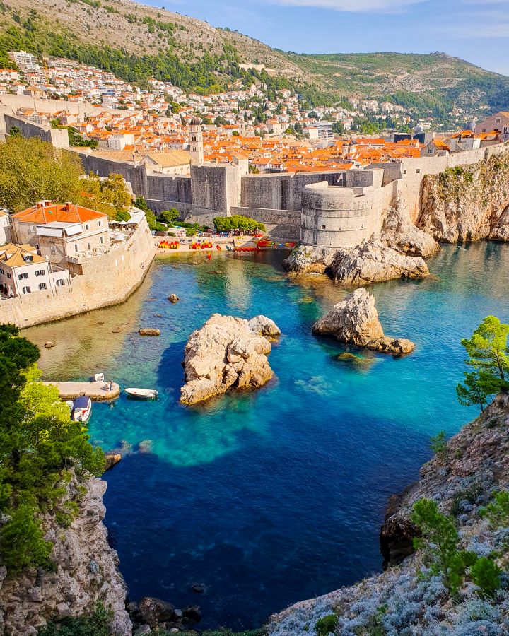 A view from Fort Lovrijenac in Dubrovnik, Croatia. In the foreground are white rocks, green trees and clear blue water. Behind the water are the Dubrovnik walls and all the orange buildings with their orange rooves. Behind the city are hills with green trees on them. "How to Spend Three Days in Dubrovnik: Best Itinerary For a First Visit"